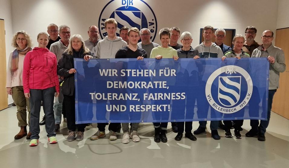 Gruppe von Menschen hält ein Banner mit der Aufschrift: „WIR STEHEN FÜR DEMOKRATIE, TOLERANZ, FAIRNESS UND RESPEKT!“